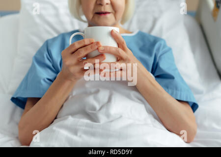 cropped view of senior woman lying in bed and drinking tea in hospital Stock Photo