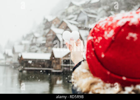 Young blonde woman in a red hat holding snow ball in a shape of a heart in front of the old village of Hallstatt, Austria. Blurry background, shallow  Stock Photo