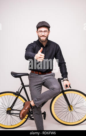 Portrait of smiling man wearing in eyeglasses and hat leaning on bicycle with thumbs up gesture over white background Stock Photo