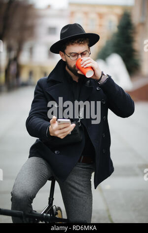 Happy young stylish man holding mobile phone while drinking coffee outdoors with bicycle Stock Photo