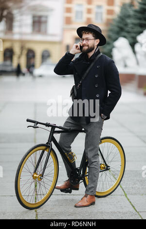 Happy young bearded man talking on the mobile phone and smiling while sitting near his bicycle outdoors Stock Photo