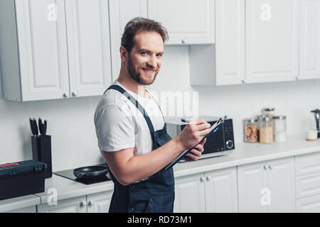 happy adult bearded repairman making notes in clipboard in kitchen Stock Photo