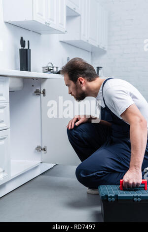 focused bearded repairman with toolbox checking sink in kitchen Stock Photo