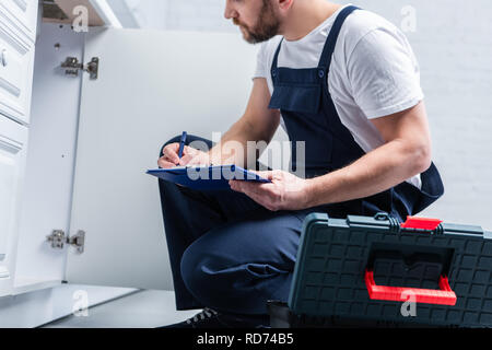 partial view of repairman in working overall writing in clipboard and checking sink in kitchen Stock Photo