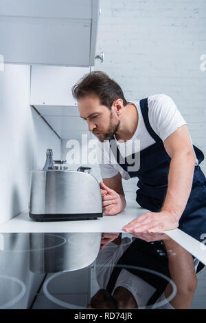 concentrated bearded craftsman in working overall fixing toaster in kitchen Stock Photo