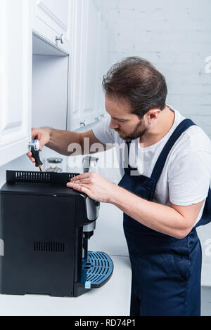 adult handyman in working overall repairing coffee machine by screwdriver in kitchen Stock Photo
