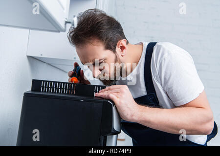 focused handyman in working overall repairing coffee machine by screwdriver in kitchen Stock Photo
