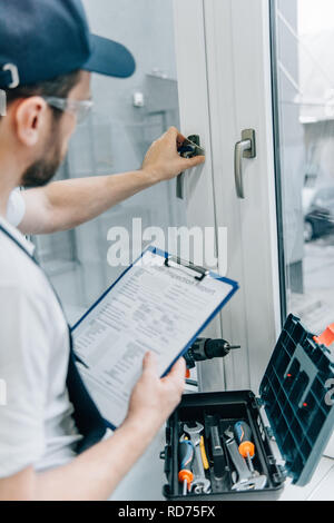 partial view of handyman in goggles holding clipboard and checking window handle Stock Photo