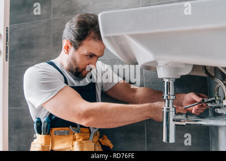 focused male plumber in working overall fixing sink in bathroom Stock Photo