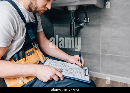 cropped shot of male plumber with toolbelt writing in clipboard near broken sink in bathroom Stock Photo