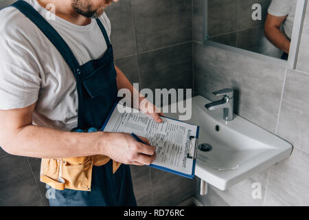 partial view of male plumber with toolbelt writing in clipboard near broken sink in bathroom Stock Photo