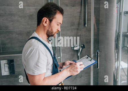 side view of male plumber making notes in clipboard while checking shower in bathroom Stock Photo