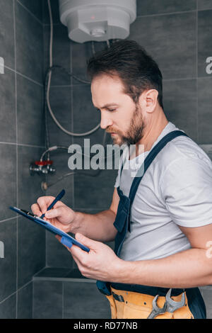 adult male plumber writing in clipboard and checking electric boiler in bathroom Stock Photo