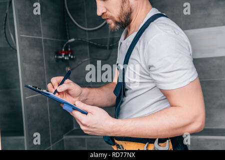 partial view of plumber writing in clipboard and checking electric boiler in bathroom Stock Photo