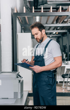 focused male craftsman checking copy machine and writing in clipboard in modern office Stock Photo