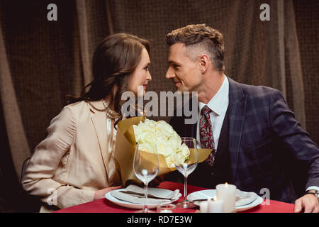 beautiful couple sitting at table with flower bouquet and looking at each other during romantic date in restaurant Stock Photo