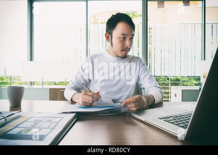 Asian smart businessman manager with casual doing his business work with laptop and business report and sitting on chair in manager room. Stock Photo