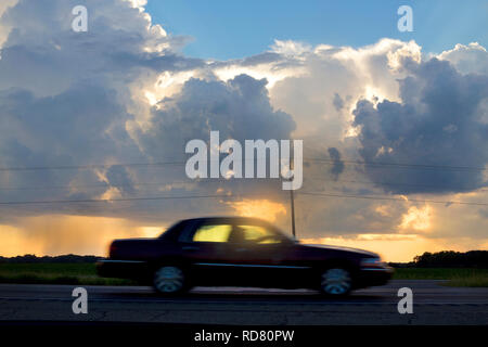 A car driving by on a rural highway with storm clouds and a sunset in the background. Stock Photo