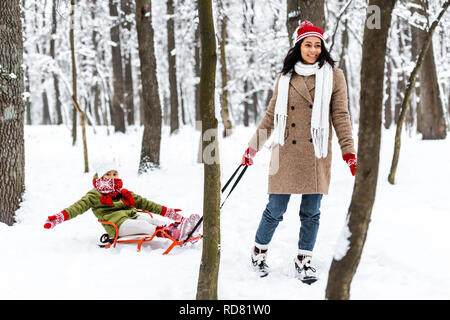beautiful african american woman smiling and pulling preteen daughter in warm clothing on sledge in winter park Stock Photo