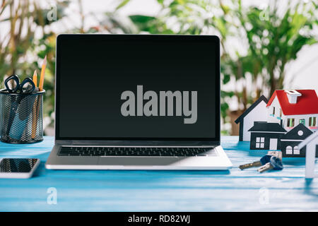 selective focus of smartphone, laptop with blank screen and keys near paper houses on wooden desk, mortgage concept Stock Photo