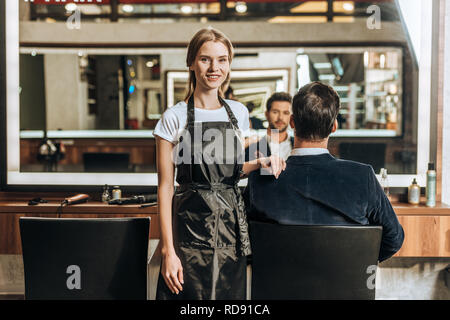 beautiful young hairstylist smiling at camera while male client sitting in chair at beauty salon Stock Photo