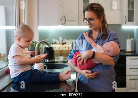 Young mother holds little daughter. Eldest son is sitting barefoot on table in kitchen and is holding an electronic thermometer. Family time. Stock Photo