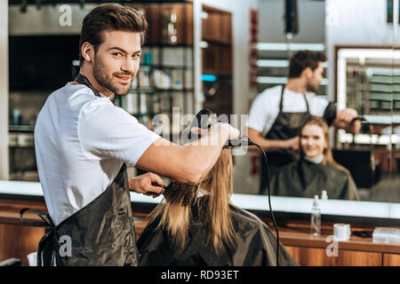 handsome young hairstylist smiling at camera while drying hair to beautiful young woman in beauty salon Stock Photo