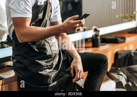 cropped shot of young male hairdresser using smartphone in beauty salon Stock Photo