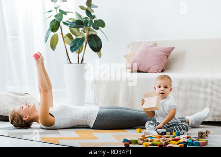 attractive woman lying on carpet and doing exercise with dumbbells and cute toddler boy holding wooden box in living room Stock Photo