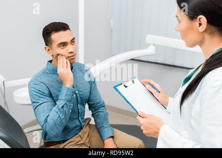 female dentist standing with clipboard and looking at handsome african american patient having toothache Stock Photo