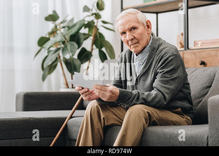 upset pensioner with grey hair holding photos while sitting on sofa Stock Photo