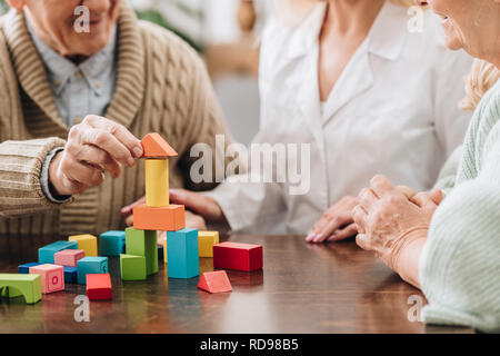 cropped view of caregiver sitting with retired man and woman and playing with wooden toys