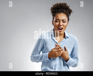 Woman holds hands on painful chest. Photo of african american woman in blue shirt on gray background. Medical concept. Heart attack. Stock Photo