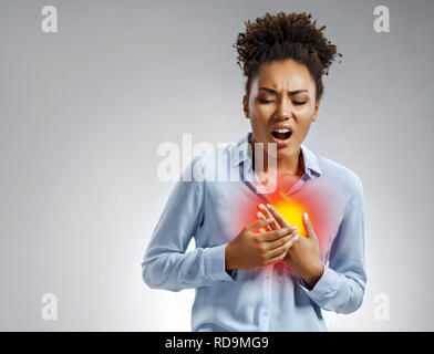 Woman holds hands on painful chest. Photo of african american woman in blue shirt on gray background. Medical concept. Heart attack. Stock Photo