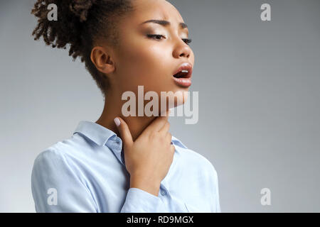 Throat pain. Woman holding her inflamed throat. Photo of african american woman in blue shirt on gray background. Medical concept Stock Photo