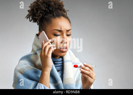 Sick woman wrapped in blanket with a high temperature calling on the phone. Photo of african american woman suffering cold and winter flu virus on gra Stock Photo