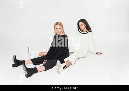 attractive multicultural friends in black and white clothes sitting and looking at camera on white Stock Photo