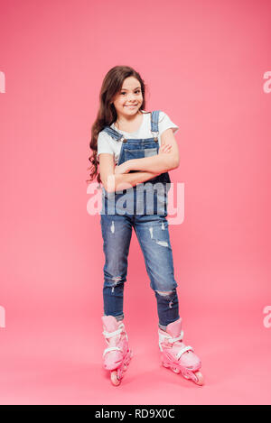 happy child in overalls with arms crossed wearing rollerblades on pink background Stock Photo