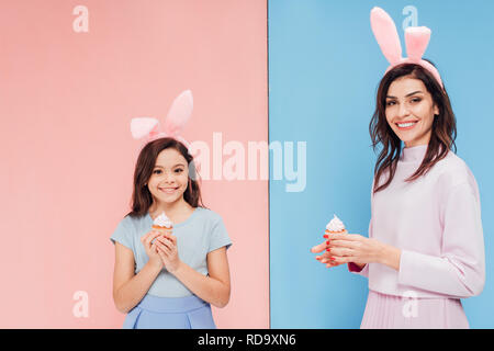 beautiful woman and child in bunny ears holding cupcakes and looking at camera on blue and pink background Stock Photo