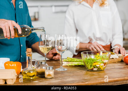 cropped image of mature husband pouring wine in glasses, wife cutting vegetables in kitchen Stock Photo