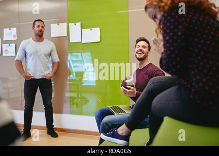 Laughing young man holding a football while sitting with colleagues during a casual meeting in a modern office Stock Photo