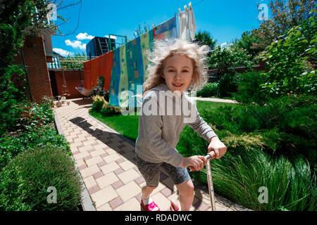 Little cheerful blonde girl riding a scooter on paved paths on a sunny summer day. Stock Photo