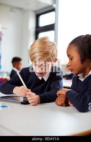 Two primary school kids using a tablet computer and stylus sitting together at desk in a classroom, vertical Stock Photo