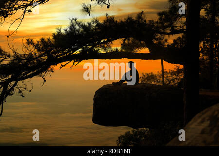 Silhouette of a man sitting on sharp rock peak with sunset background. Stock Photo