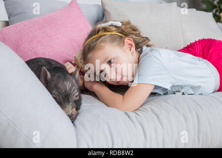 Beautiful baby girl on sofa near a black vietnamese piglet. Concept of the Chinese New Year 2019 of the Pig.Mini piggy,pig, symbol of 2019 in the Chinese calendar.Winter holidays concept. Stock Photo