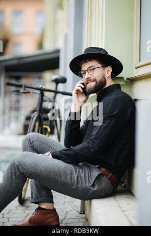 Handsome bearded businessman in classic suit is using a smart phone and smiling while riding bicycle in city Stock Photo
