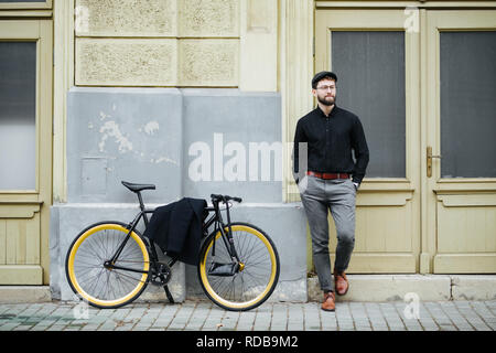 Full length portrait of handsome young man in casual clothes looking away and smiling while leaning near bike, standing outdoors Stock Photo