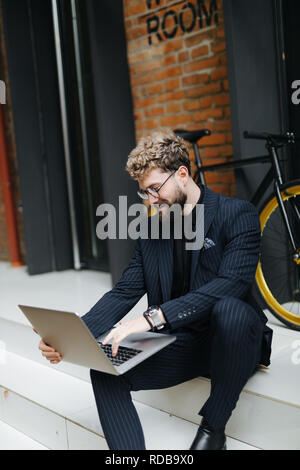 Attractive young bearded man working with laptop sitting on the floor in the street with bicycle Stock Photo