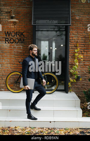 Attractive young man walking down stairs with laptop in hands with vintage bicycle on background in the street Stock Photo