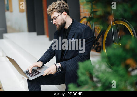 Attractive young bearded man working with laptop sitting on the floor in the street with bicycle Stock Photo